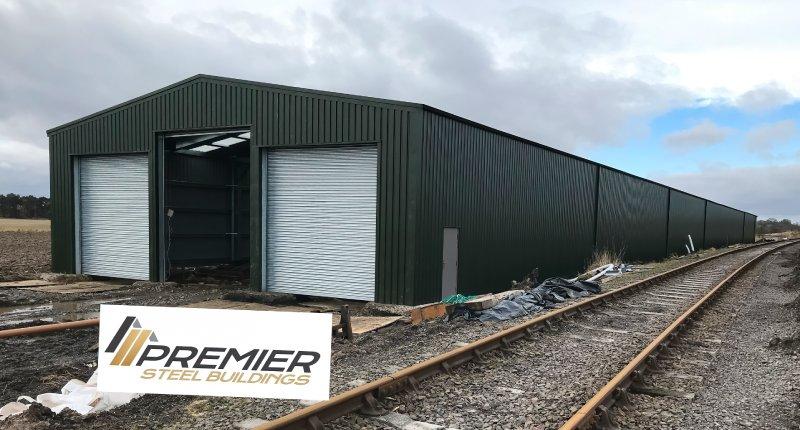 Perspective view of a long olive green industrial steel building situated next to railway tracks, showing the side profile and front entrance bays.